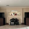A living room showcases Vista 3-Way Horn Speakers flanking the fireplace, with a shelving unit for audio gear. The mantel is adorned with vases and red flowers. A living room showcases Vista 3-Way Horn Speakers flanking the fireplace, with a shelving unit for audio gear. The mantel is adorned with vases and red flowers.