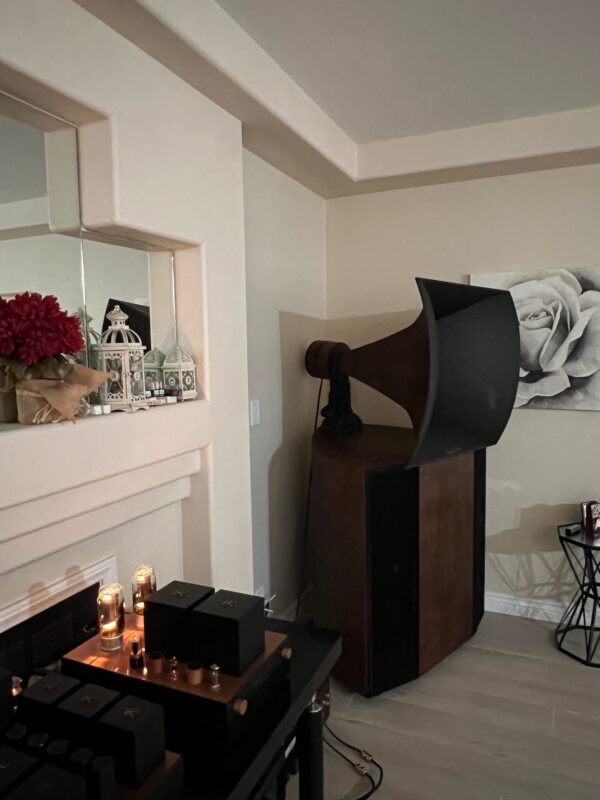 A Vista 3-Way Horn Speaker with a vintage flair stands by the wall adorned with a rose painting. Nearby, electronic audio components are placed on a stand alongside decorative items on the mantel. A Vista 3-Way Horn Speaker with a vintage flair stands by the wall adorned with a rose painting. Nearby, electronic audio components are placed on a stand alongside decorative items on the mantel.