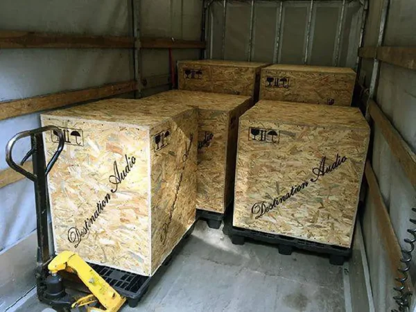 Three large wooden crates labeled "Vista 3-Way Horn Speakers" inside a shipping truck, arranged with a pallet jack nearby. Three large wooden crates labeled "Vista 3-Way Horn Speakers" inside a shipping truck, arranged with a pallet jack nearby.